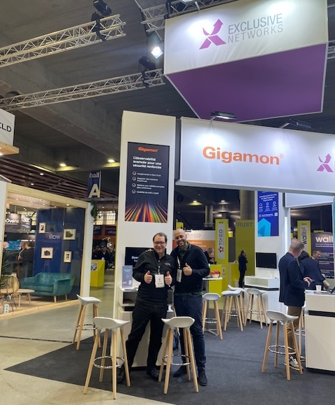 Two individuals posing with thumbs up at a trade show booth for Exclusive Networks and Gigamon. The booth features modern furniture, branded banners, and overhead signage with both company logos. The environment is vibrant, with other booths and attendees visible in the background, highlighting a professional networking event.